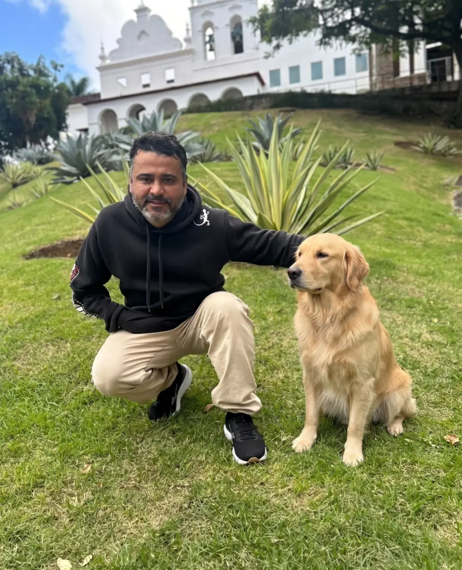 V. Júnior agachado ao lado de um golden retriever sentado e obediente, em área verde da Grande Vitória
